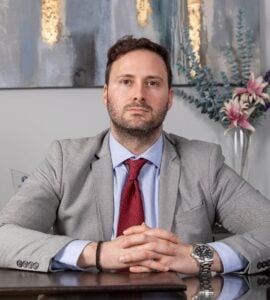 Attorney seated at desk with hands clasped, dressed in a grey suit and red tie, in a modern office setting.