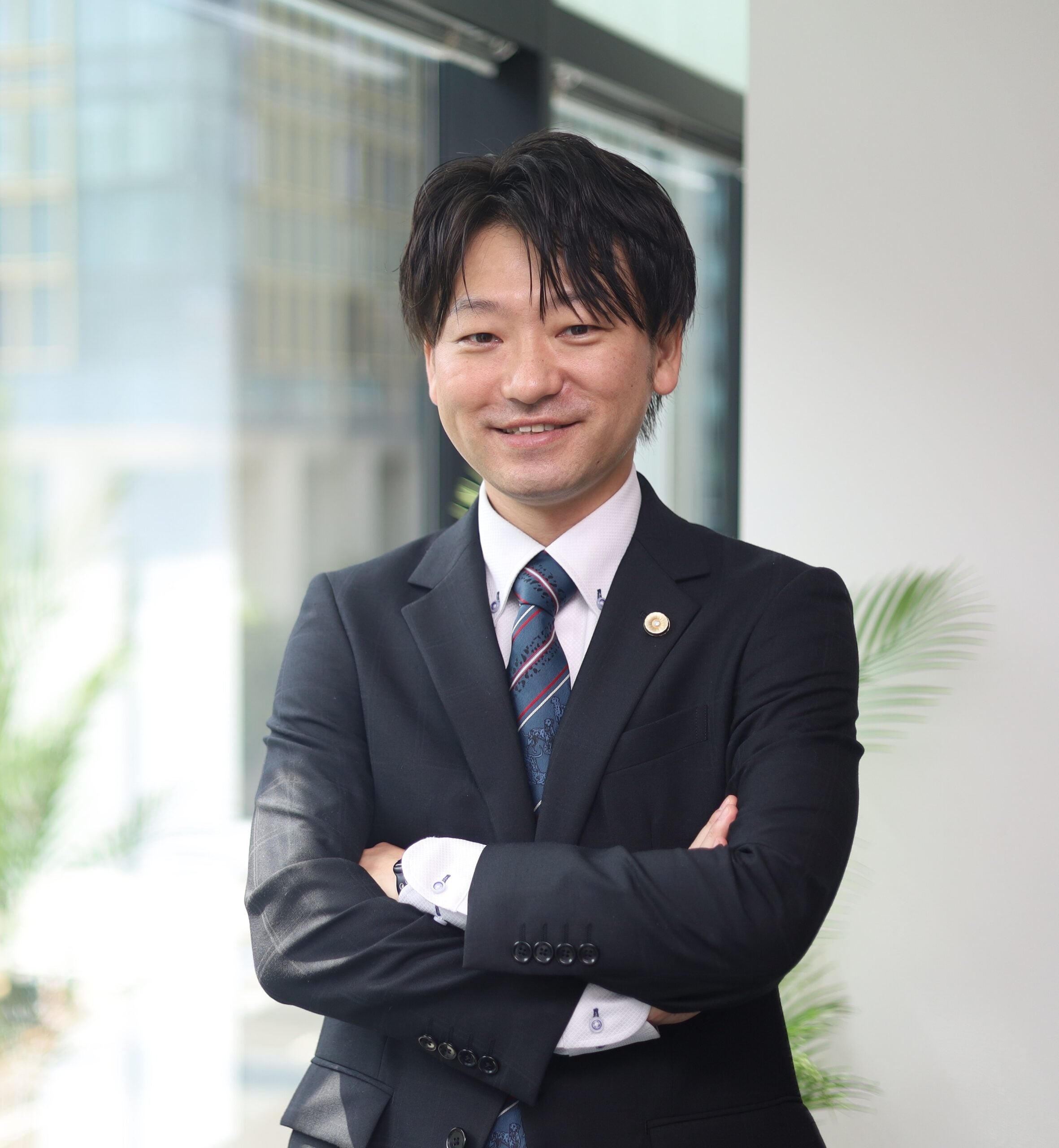 Attorney smiling while standing with arms crossed in a modern office setting with windows and greenery in the background.