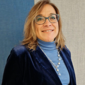 Professional woman smiling, wearing glasses and a blue blazer, in a neutral indoor setting.