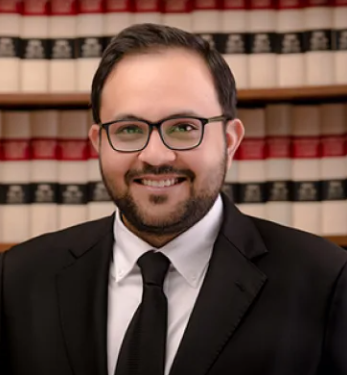 Lawyer smiling in front of shelves filled with law books, dressed in a black suit and glasses.
