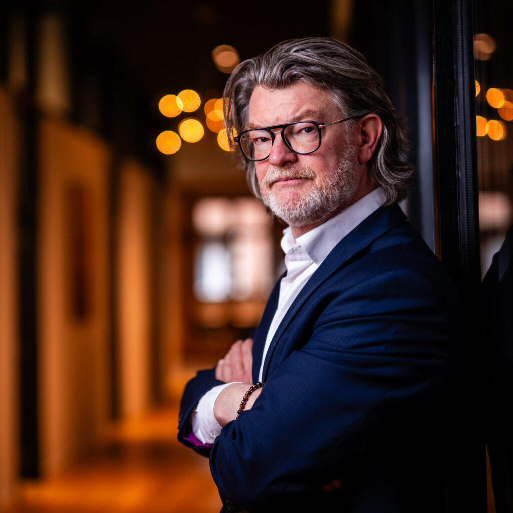 Senior lawyer posing confidently with arms crossed in a well-lit office hallway.