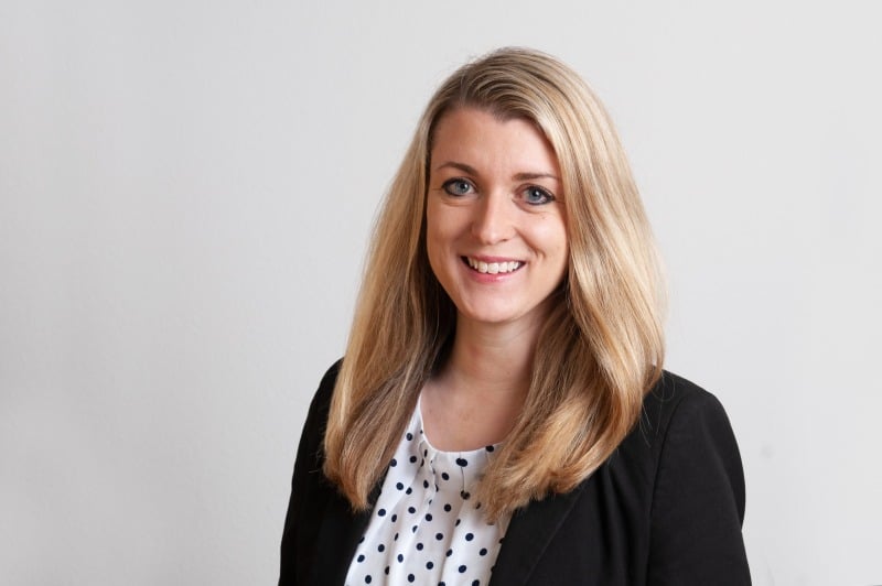 Attorney with long blonde hair smiling at the camera, wearing a black blazer and white blouse against a light background.