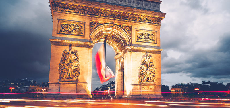 Arc de Triomphe illuminated at night with a flag hanging from the arch, surrounded by city traffic and dramatic clouds.