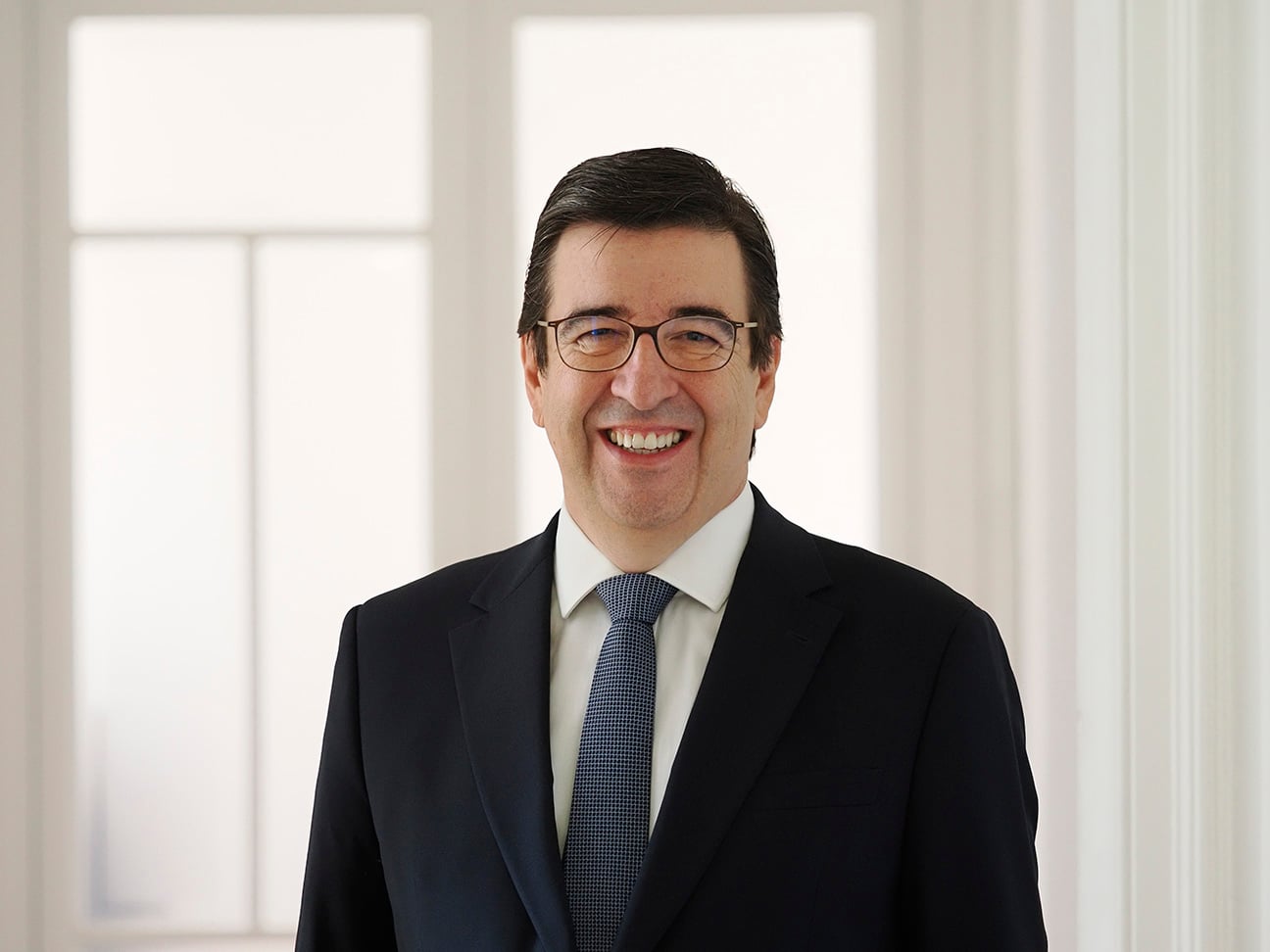 Lawyer smiling in a professional suit, standing in a bright office setting with large windows.