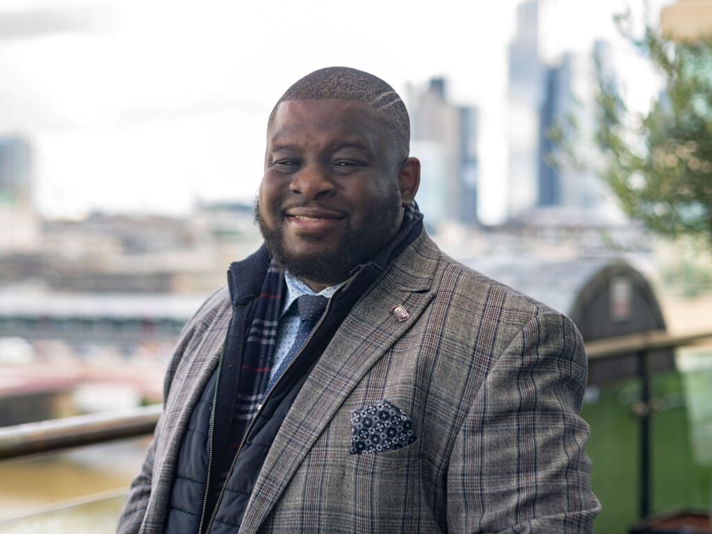 Smiling professional wearing a checked suit, standing outdoors with a city skyline in the background.