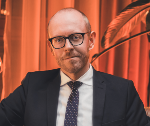 Lawyer sitting confidently, wearing a suit, in a professional environment with warm lighting and a plant in the background.