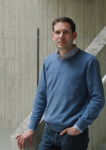 Male professional leaning against a concrete wall in a business casual setting.