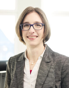 Lawyer smiling in a modern office setting, wearing a professional suit with short hair and glasses.