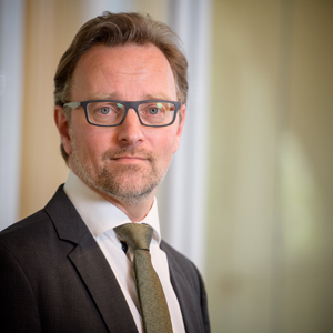 Lawyer in suit and glasses, standing in well-lit office environment, looking directly at the camera.