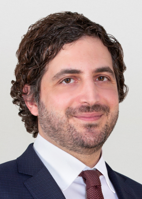 Lawyer with curly hair wearing a dark suit and tie, smiling against a neutral background.
