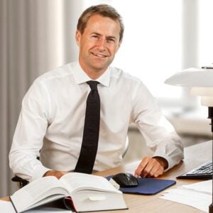 Lawyer sitting at desk with open legal book and computer, smiling in a bright office setting.