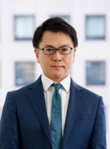 Attorney wearing glasses in a blue suit, standing in a modern office with large windows in the background.