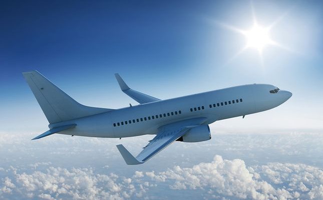 Commercial airplane flying in clear blue sky above fluffy white clouds with sunlight.