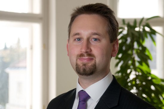 Professional man in a suit standing near a window with greenery in an office setting.