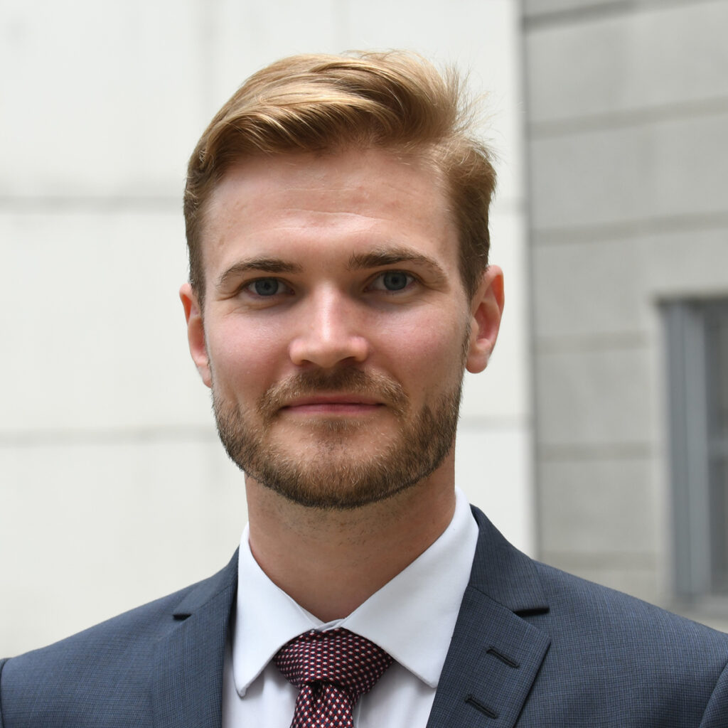 Young man in a suit and tie, standing outdoors near a legal building, looking confidently at the camera.