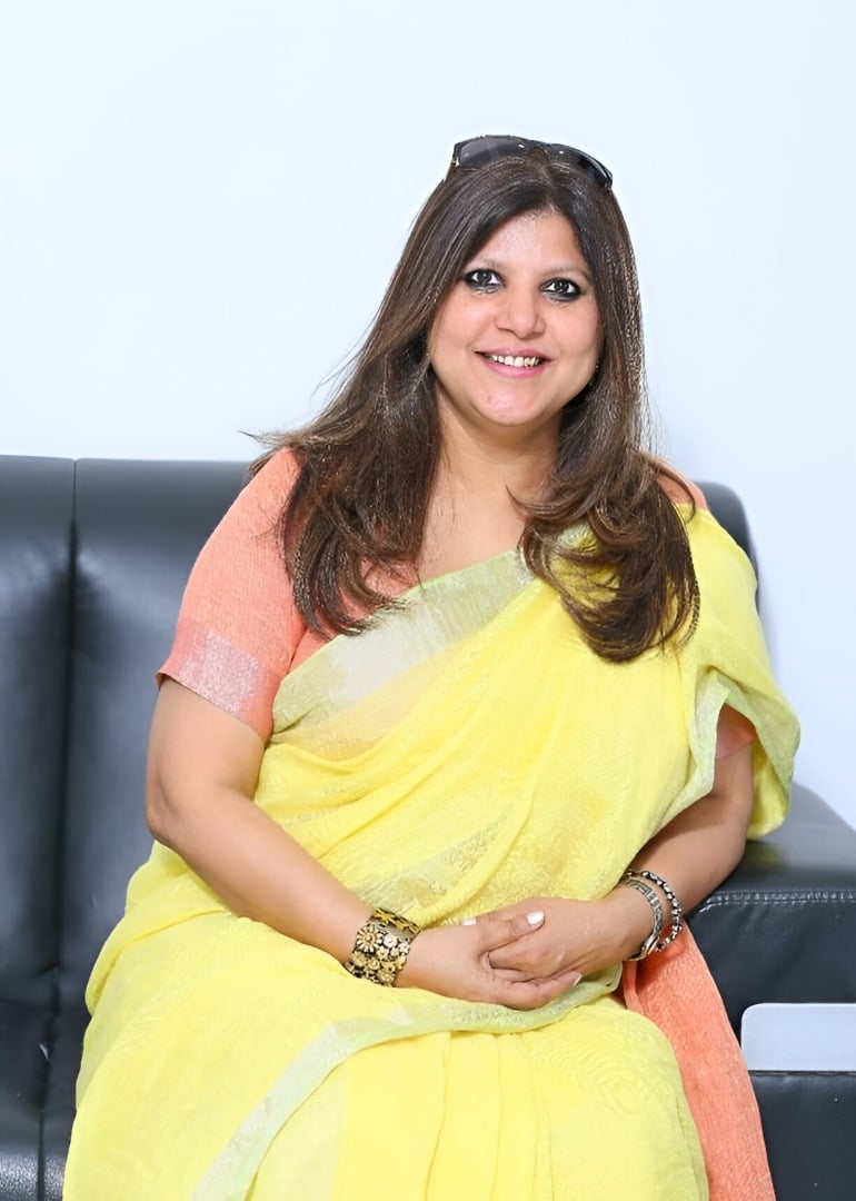 Legal professional seated in a black chair, wearing a yellow sari, with a neutral expression in a well-lit setting.