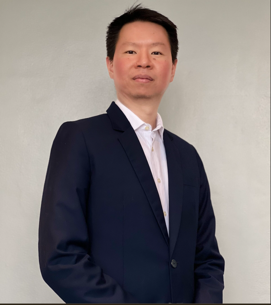 Professional individual in a dark suit standing against a neutral background, conveying a confident demeanor.