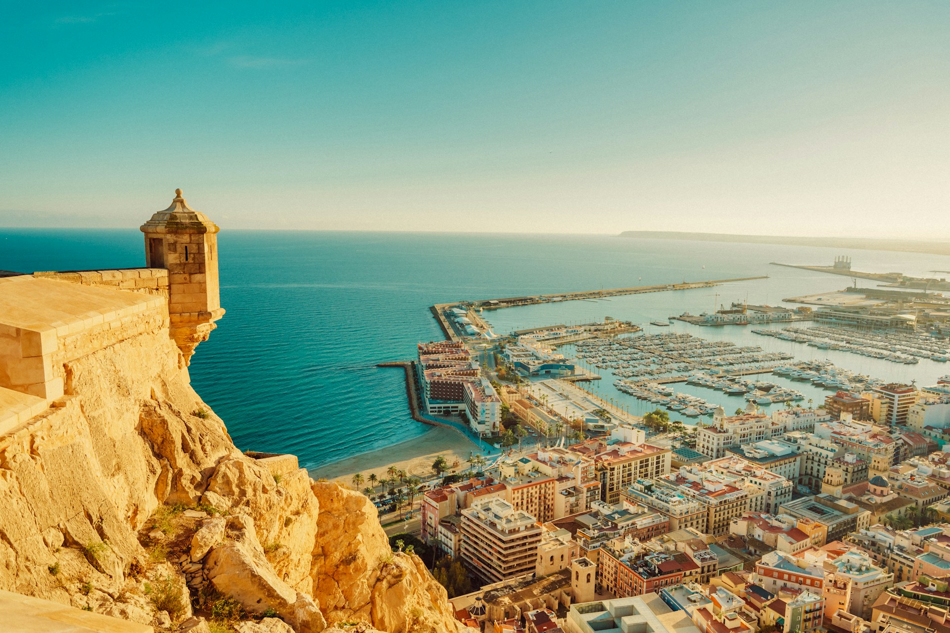 Coastal view of a city with a marina and beach, featuring buildings and a lighthouse on the cliff.