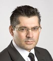 Professional male lawyer with short black hair and glasses, dressed in a suit against a neutral background.