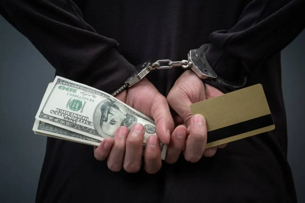 Person in handcuffs holding cash and a credit card, indicating a potential legal situation involving theft or fraud.