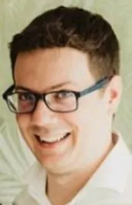 Man with glasses smiling, against a light background, appears in a casual setting.