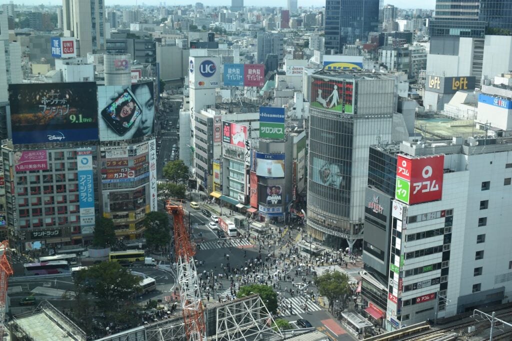 Busy urban intersection filled with pedestrians and vehicles, surrounded by tall commercial buildings and advertisements.