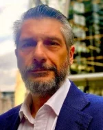 Professional man with a beard standing outdoors in smart attire, likely associated with a law or business environment.