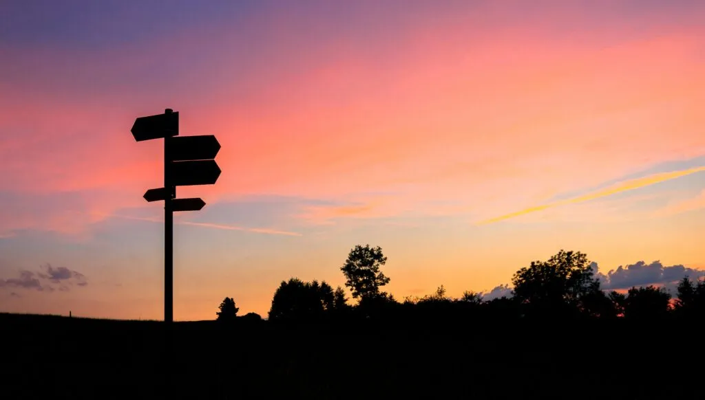 Silhouette of a signpost against a colorful sunset sky with trees in the background.