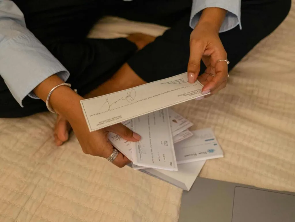 Individual holding envelopes and checks while seated on a bed, with documents and a laptop nearby.