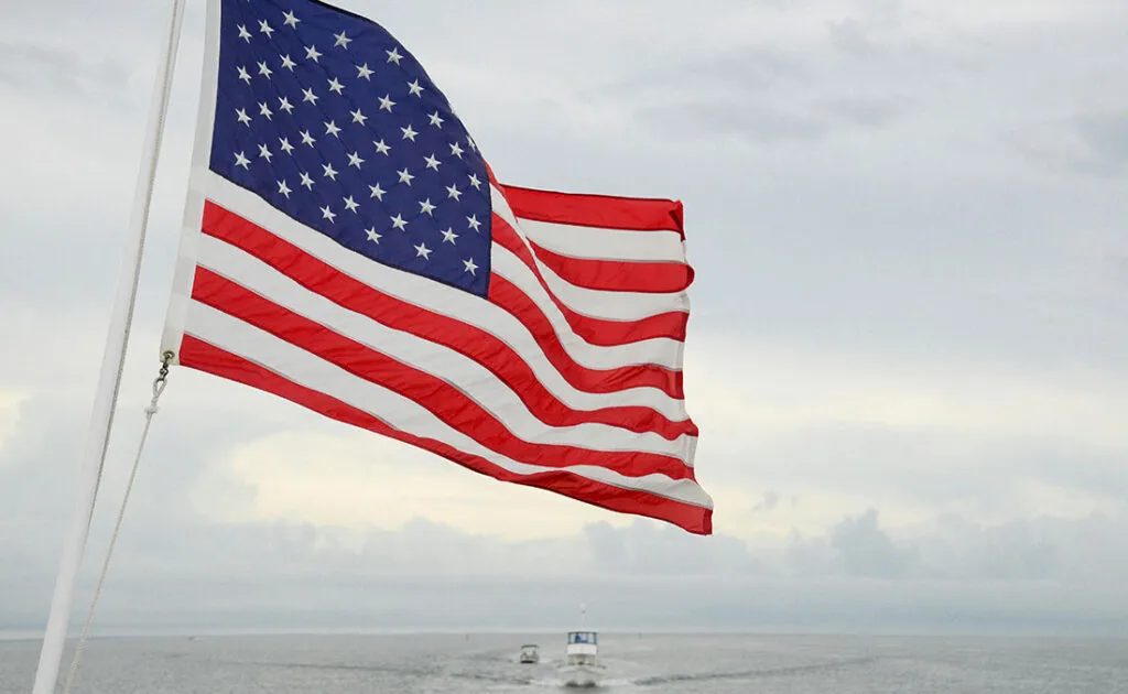 American flag waving against a cloudy sky, with a boat visible in the distance on the water.