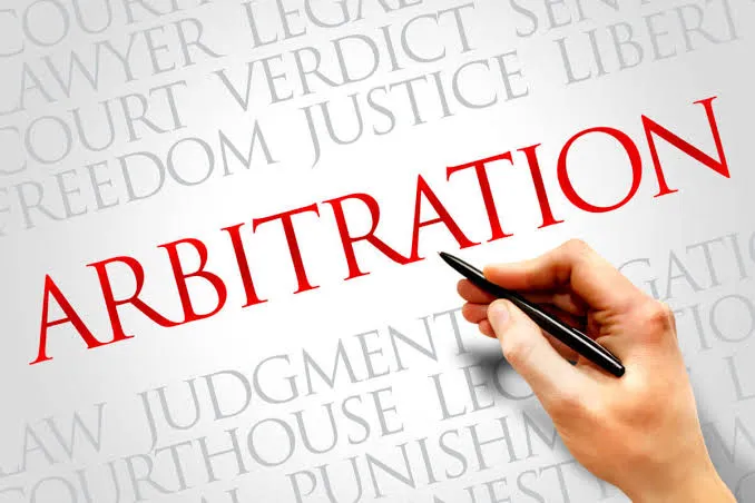 Hand holding a pen writing "ARBITRATION" with legal terms like "law," "court," and "justice" in the background.