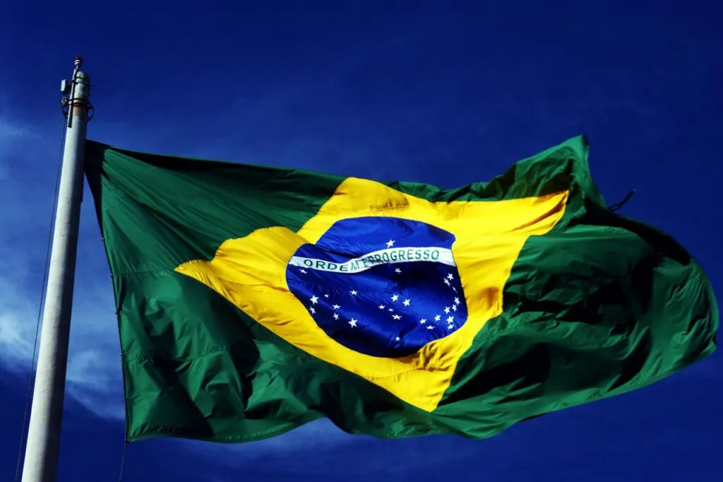 Brazilian flag waving against a blue sky, featuring green and yellow colors with a blue globe and white stars in the center.
