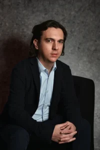 Professional man seated on a chair, dressed in a blazer and shirt, posing in a neutral-toned setting.