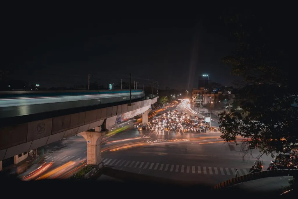 Nighttime urban scene with traffic lights illuminating busy intersection and an elevated train track above.