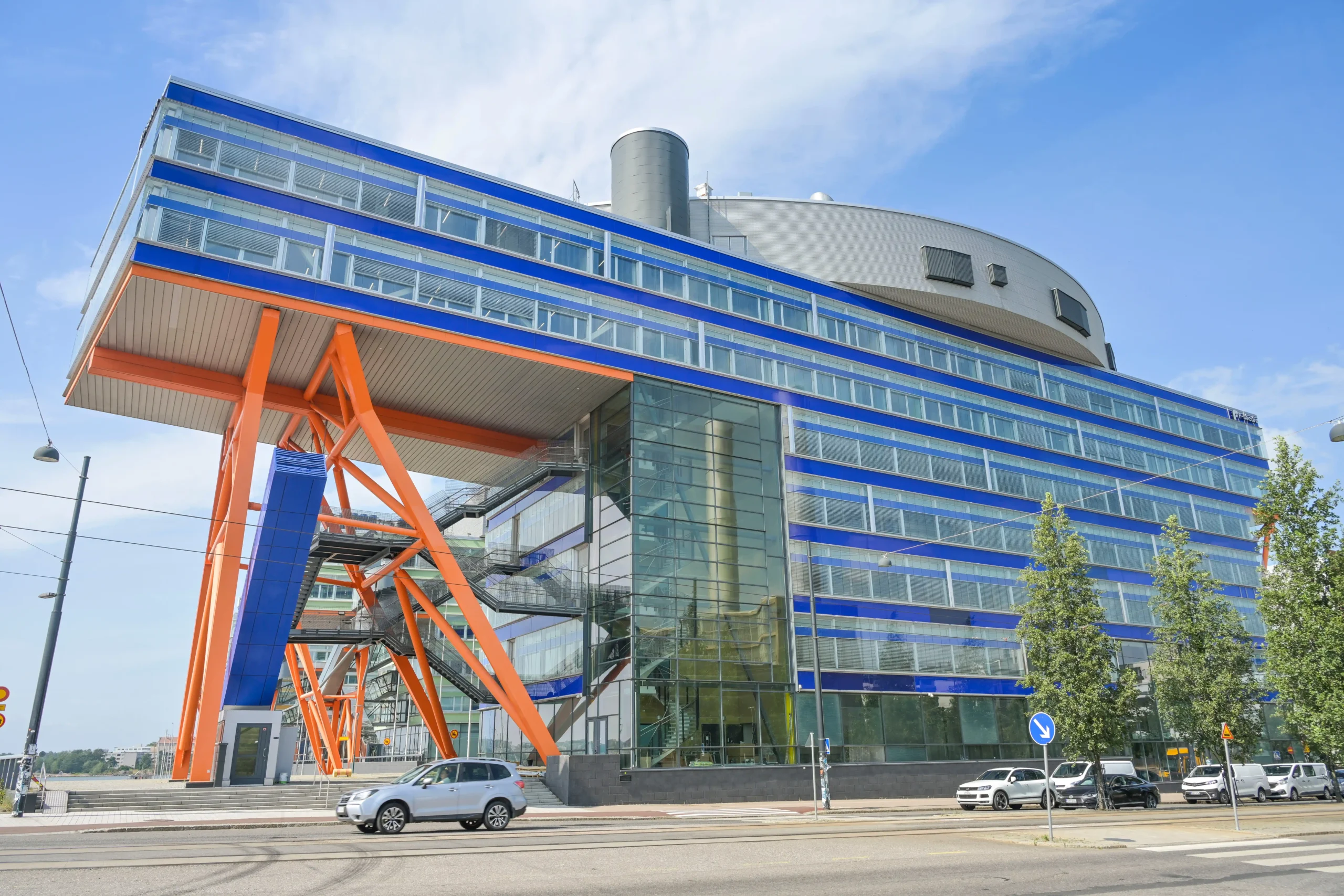 Modern office building with blue and orange accents, featuring large glass windows and adjacent visitor parking area.